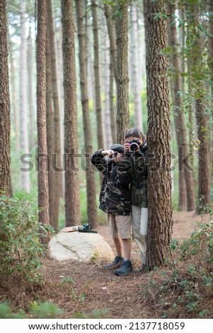 Happy mother and son spending time in forest. Woman and son in casual clothes with cameras peeking from behind trees. Hobby, family, nature, photography concept