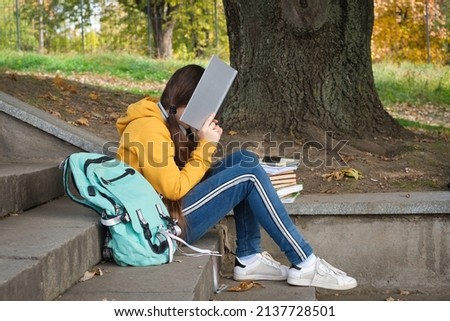 Tired teenage girl reads a book, covers her face with a book sitting on the stairs in the park.