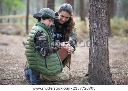 Family of amateur photographers in forest. Woman and son in casual clothes with cameras kneeling near tree. Hobby, family, nature, photography concept