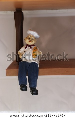 Porcelain chef doll on a shelf. Kitchen decoration in the closet. A chef doll holds bread and a knife. A doll in a traditional chef costume. Shelf doll. An ornamental object on a shelf hanging under
