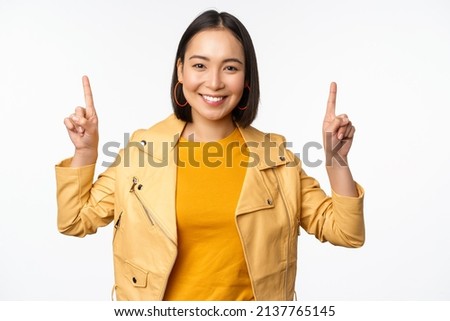 Enthusiastic asian girl pointing fingers up, showing advertisement on top, smiling happy, demonstrating promo offer or banner, standing over white background