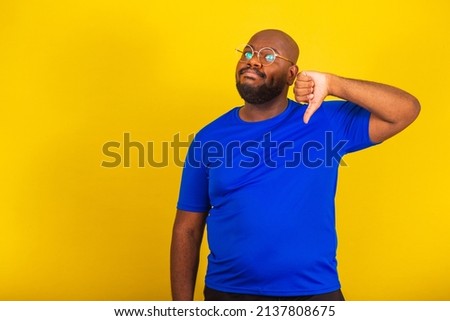 handsome afro brazilian man wearing glasses, blue shirt over yellow background. thumbs down, disapproving, disappointment, disappointed, disapproving, negative.