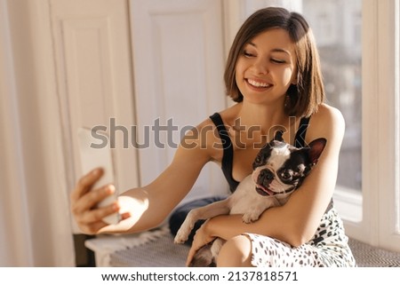 Joyful caucasian young girl takes picture of herself on front camera of smartphone with dog. Brunette wears summer clothes while sitting on windowsill at home. Pets and technology concept