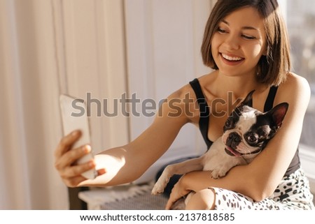 Young caucasian lady takes selfie with dog using smartphone camera while sitting on windowsill. Brunette having fun with cute animal. People and technology concept.