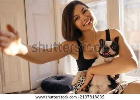 Portrait of smiling young caucasian woman holding boston terrier and taking selfie photo. Brunette with short haircut wears summer clothes. Love for pets, joy and tenderness