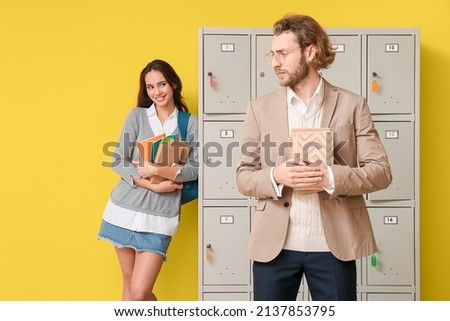 Female student looking at her teacher while standing near locker on yellow background