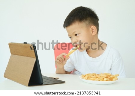Asian 5 years old little kindergarten boy child eating French fries while watching a video from tablet pc isolated over white wall background, Unhealthy Kids Foods, Gadget-addicted children concept