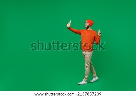 Full body young fun smiling happy caucasian man 20s wear orange sweatshirt hat doing selfie shot on mobile cell phone post photo on social network show v-sign isolated on plain green background studio