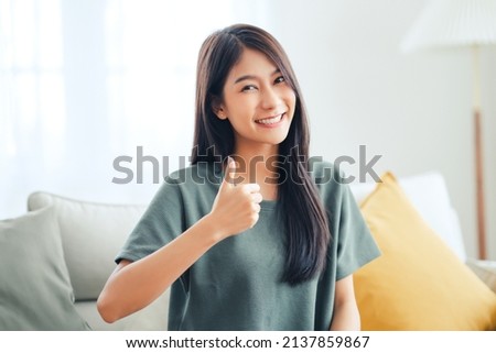 Satisfied asian woman looking at camera with thumbs up sitting on a couch in the living room at home. Royalty-Free Stock Photo #2137859867