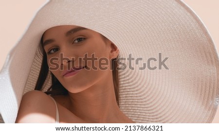 Young gorgeous dark-haired European woman in a big white hat with a smear of sunscreen on her cheek lifts up her shoulder and looks at camera against beige background | Sunscreen concept Royalty-Free Stock Photo #2137866321