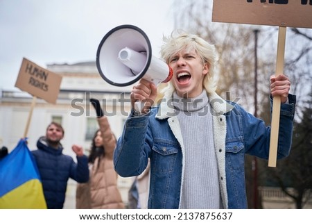 Caucasian man  screaming through megaphone in front and group of people manifesting  in the background