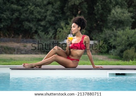 A young African American woman in a red bikini sunbathes by the pool while sipping a non-alcoholic fruit cocktail - gen z people in luxury vacation lifestyle
