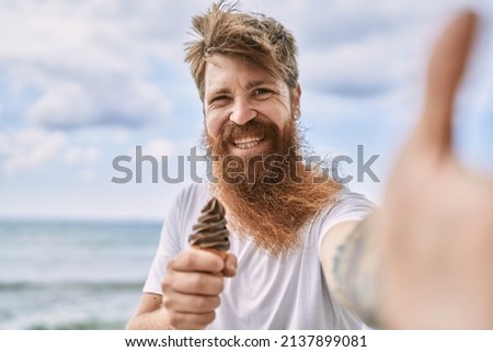 Young redhead man eating ice cream make selfie by the camera at the beach.