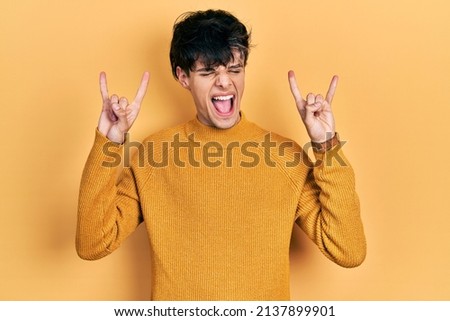Handsome hipster young man wearing casual yellow sweater shouting with crazy expression doing rock symbol with hands up. music star. heavy music concept. 