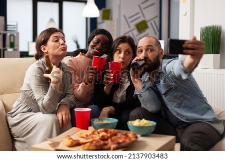Group of people taking selfies and celebrating with drinks in office after work hours. Man and women having fun with pictures on smartphone, enjoying alcoholic drinks and snacks at party.
