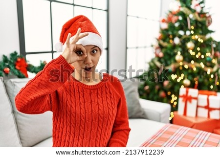 Young hispanic woman with short hair wearing christmas hat sitting on the sofa doing ok gesture shocked with surprised face, eye looking through fingers. unbelieving expression. 