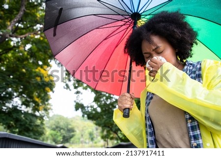 Front view portrait of an African American woman walking towards camera sneezing holding an colorful umbrella under the tree at public park. Health care and insurance concept