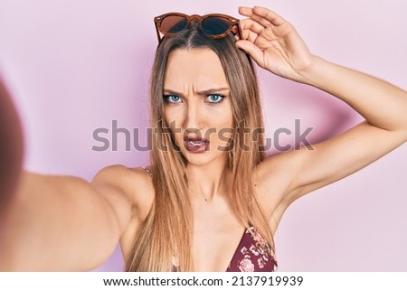 Young blonde girl wearing bikini taking a selfie in shock face, looking skeptical and sarcastic, surprised with open mouth 