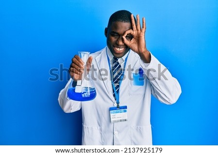 Young african american man wearing scientist uniform holding test tube smiling happy doing ok sign with hand on eye looking through fingers 