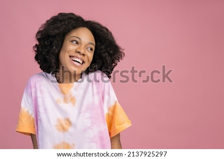 Beautiful smiling African woman looking away isolated on pink background, copy space. Happy curly haired fashion model wearing stylish tie dye t shirt posing for pictures in studio 