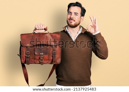 Young hispanic man wearing leather bag doing ok sign with fingers, smiling friendly gesturing excellent symbol 