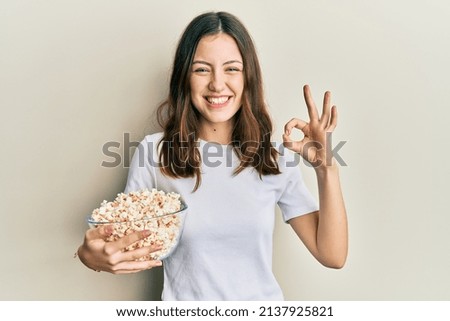 Young brunette woman eating popcorn doing ok sign with fingers, smiling friendly gesturing excellent symbol 