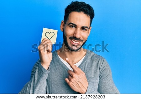 Young hispanic man holding heart reminder smiling happy pointing with hand and finger 