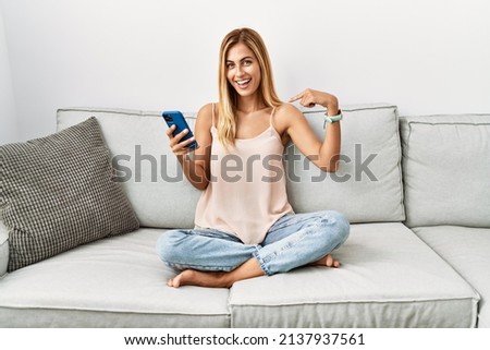 Blonde beautiful young woman sitting on the sofa at home using smartphone looking confident with smile on face, pointing oneself with fingers proud and happy. 