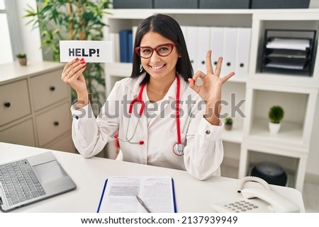 Young hispanic doctor woman holding help banner doing ok sign with fingers, smiling friendly gesturing excellent symbol 