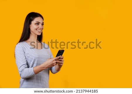 Young beautiful brunette woman with long hair wearing striped shirt, posing with a phone over isolated yellow background. Portrait of female model typing on her cellphone. Close up, copy space.