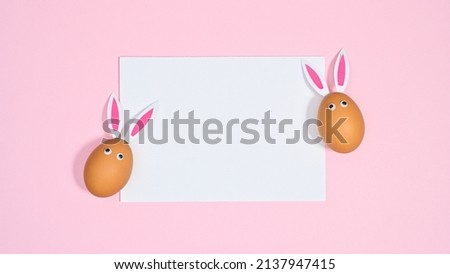 Two natural eggs with bunny ears and eyes on pastel pink background. Flat lay copy space paper card Easter concept