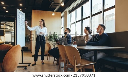 Female manager giving a presentation to her team during a meeting in a modern workspace. Confident young businesswoman discussing her marketing strategy and ideas with her colleagues.