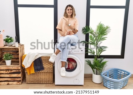 Young hispanic woman drinking coffee waiting for washing machine at laundry room cheerful with a smile on face pointing with hand and finger up to the side with happy and natural expression 