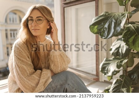 Middle-aged european woman with long blond hair looks at camera while sitting on windowsill near open window. Model wears glasses, sweater and jeans. Lifestyle, leisure concept. Royalty-Free Stock Photo #2137959223