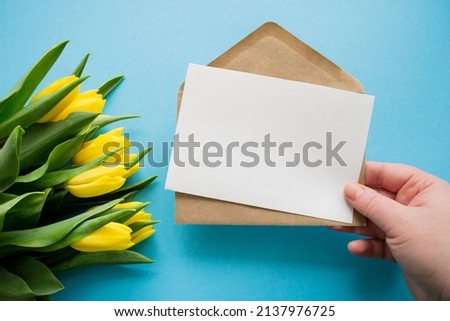 Hand holds envelope and paper for text on blue background and yellow tulips. Happy holiday greetings