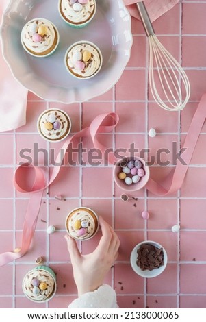 Flat lay Easter baking concept image. Easter theme cupcakes image with chocolate and baking equipment included in frame. Woman's hand reaches into frame to take a frosted cup cake. Pastel pink colours