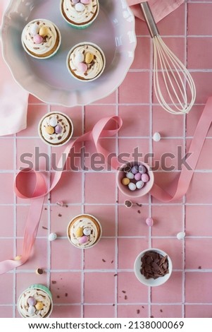 Flat lay Easter baking concept image. Easter theme cupcakes image with chocolate and baking equipment included in frame. Set against a pink tile background. Light pastel pink and cream colour theme