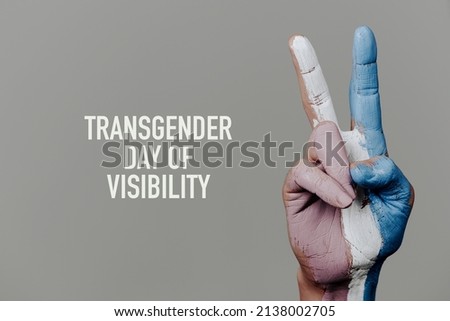 the text transgender day of visibility and the hand of a person doing the V-sign with the transgender pride flag painted in it, on a pale gray background Royalty-Free Stock Photo #2138002705