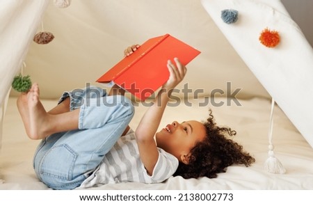 Portrait of cute little african american kid curly boy with book smiling at camera while   reading book  in play tent at home, happy child playing alone in children room Royalty-Free Stock Photo #2138002773