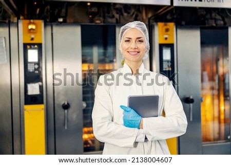 A smiling female food factory inspector with tablet in hands standing in front of ovens in bakery. Royalty-Free Stock Photo #2138004607