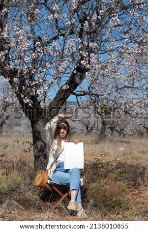 Young woman sitting under a tree with flowers and a notebook in her hand, covering the sun on her face on a spring day