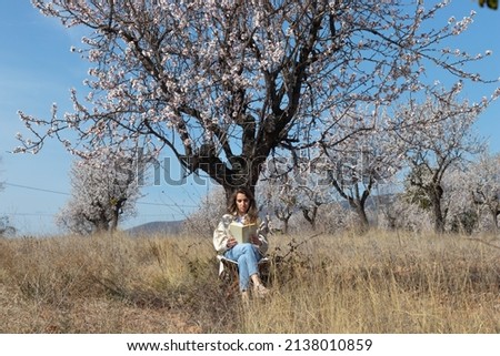 Landscape panorama with seated woman reading book under an almond tree in a field under blue sky