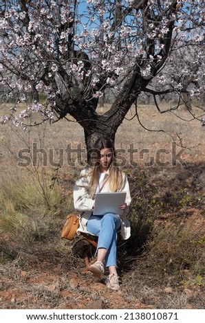 Forehead portrait of a young and beautiful woman sitting and leaning on the trunk of almond tree in bloom while drawing