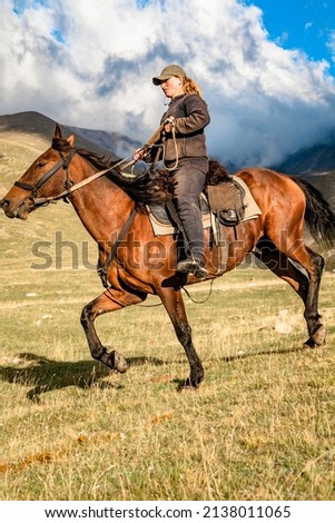 A cowgirl cowherd rider on a horse gallops through a mountain valley on a clear autumn day sky with clouds