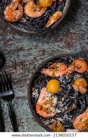 Dark ceramic plate with black shrimp spaghetti on a grey-blue textured background. A glass of white wine, cutlery, parmesan and a second serving in the background. Menu for restaurant, cafe. Top view.