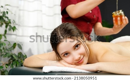 Close up photo of a young pretty blond woman lying on a massage table looking at the camera whilr masseur preparing honey for procedure.