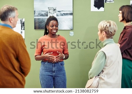 Portrait of cheerful young Black female creator standing in front of art gallery exhibition visitors speaking about photos