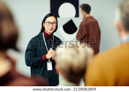 Young Asian man working in modern arts gallery standing in front of group of unrecognizable people speaking about exhibition Royalty-Free Stock Photo #2138082809