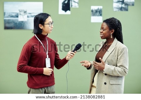Young Asian art gallery worker taking interview of modern cheerful African American creator on first exhibition day