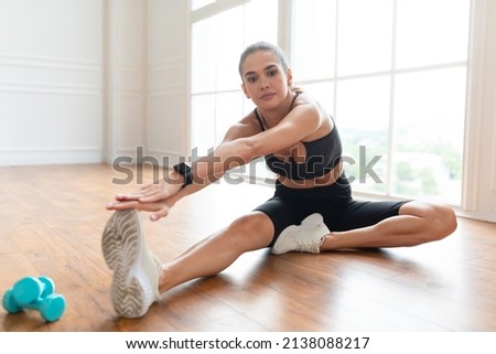 Motivation And Effort. Strong confident athletic lady in sportswear and sneakers stretching leg muscles sitting on the floor at gym or in minimal living room interior, looking at camera, copy space